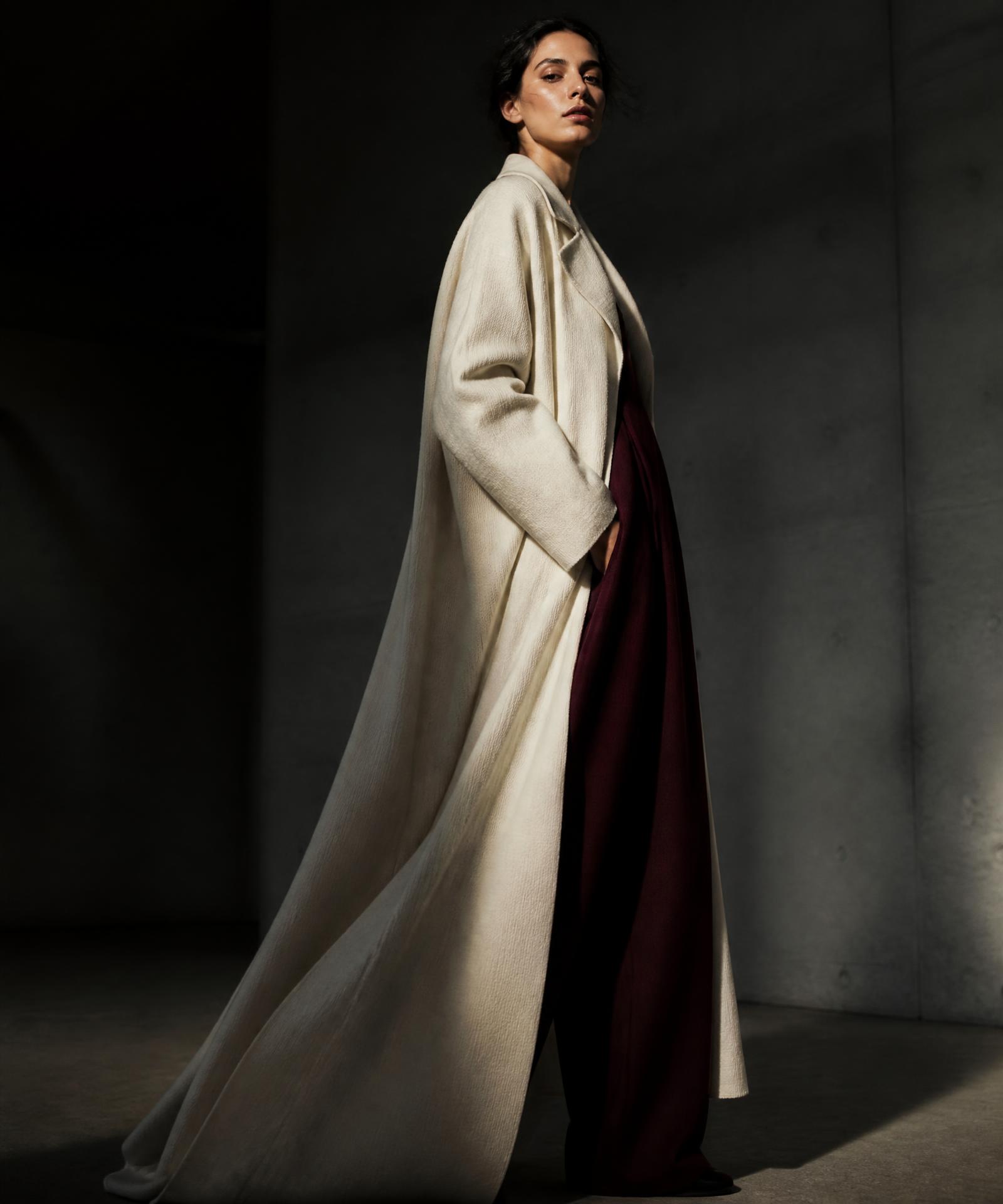 Model wearing an ivory VELORA overcoat over burgundy trousers